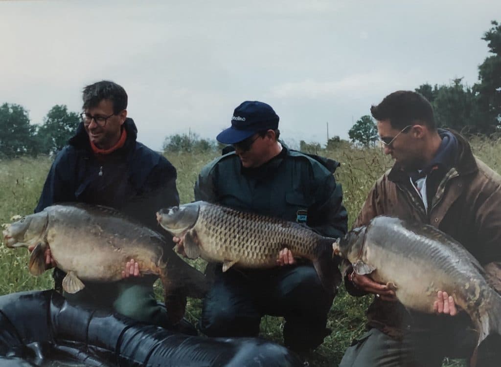 3 Frenchmen on their knees in a field, each holding a large carp fish